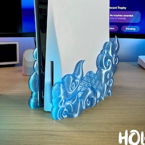 May include: A white PlayStation 5 console is displayed on a blue 3D printed stand shaped like a cloud. The stand is on a wooden surface. The text "HOLO - PROPS" is visible in the lower right corner of the image.