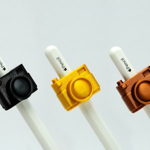 May include: Three Apple Pencil caps with camera designs. The caps are black, yellow, and copper.