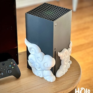 May include: A black gaming console with white cloud-shaped decorative accents. The console is on a wooden table, with a black controller visible on the left. The logo "HOLO - PROPS" is in the bottom right corner.