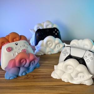 Peut inclure: Trois supports d'impression 3D en forme de nuage de différentes couleurs pour manettes de jeux vidéo. Les supports sont blancs, roses et bleus, et chacun a une manette différente posée dessus.