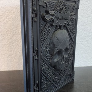 Op de afbeelding: Zwart, in reli&euml;f uitgevoerd boek met het woord "Deathnote" op de kaft. De kaft heeft een schedel en sierlijke, gotische details. Het boek staat rechtop op een donkere ondergrond.