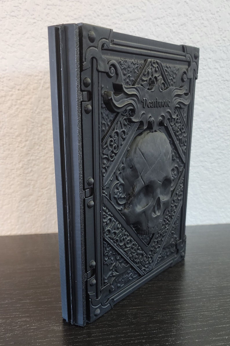 Op de afbeelding: Zwart, in reli&euml;f uitgevoerd boek met het woord "Deathnote" op de kaft. De kaft heeft een schedel en sierlijke, gotische details. Het boek staat rechtop op een donkere ondergrond.