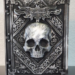 Op de afbeelding: Een gedetailleerd, grijs en zilver Death Note-boek met een schedel op de omslag. Het boek heeft sierlijke, gotische details en de titel "Deathnote" wordt prominent weergegeven. Het boek is gesloten en lijkt van een hard materiaal te zijn.