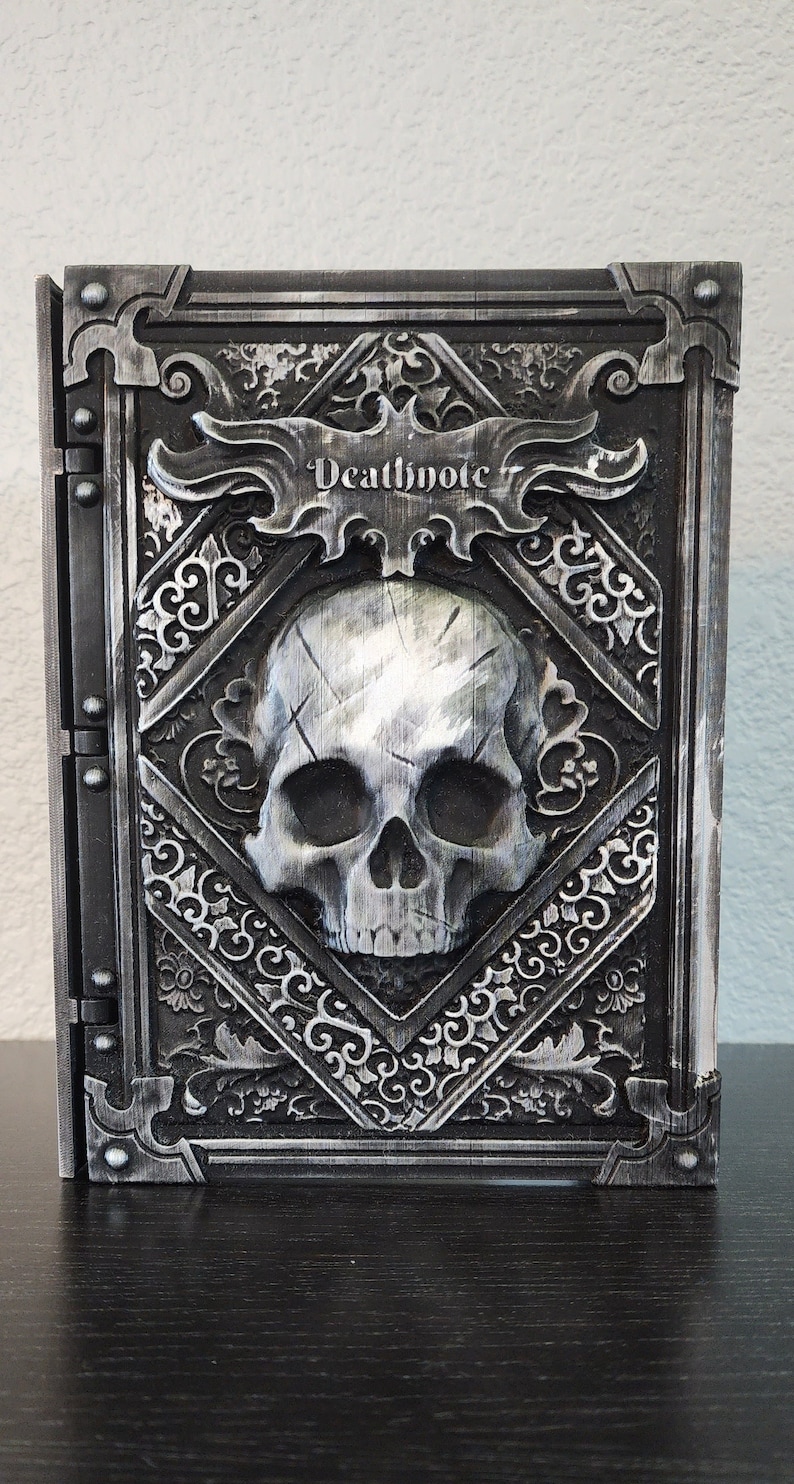Op de afbeelding: Een gedetailleerd, grijs en zilver Death Note-boek met een schedel op de omslag. Het boek heeft sierlijke, gotische details en de titel "Deathnote" wordt prominent weergegeven. Het boek is gesloten en lijkt van een hard materiaal te zijn.
