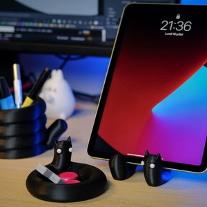 May include: A black 3D-printed desk organizer set featuring a tablet stand, pen holder, and small tray. The tablet displays a red, blue, and purple gradient with the time 21:36. The accessories have a cat-like design.