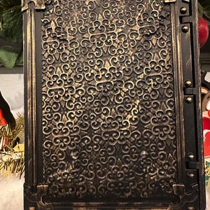 Op de afbeelding: Een rechthoekige, sierlijke, zwart-gouden decoratieve doos. De doos heeft een gedetailleerd, wervelend patroon op het voorpaneel, omlijst door een goudkleurige rand met decoratieve hoekaccenten. De doos heeft een scharnierzijde.
