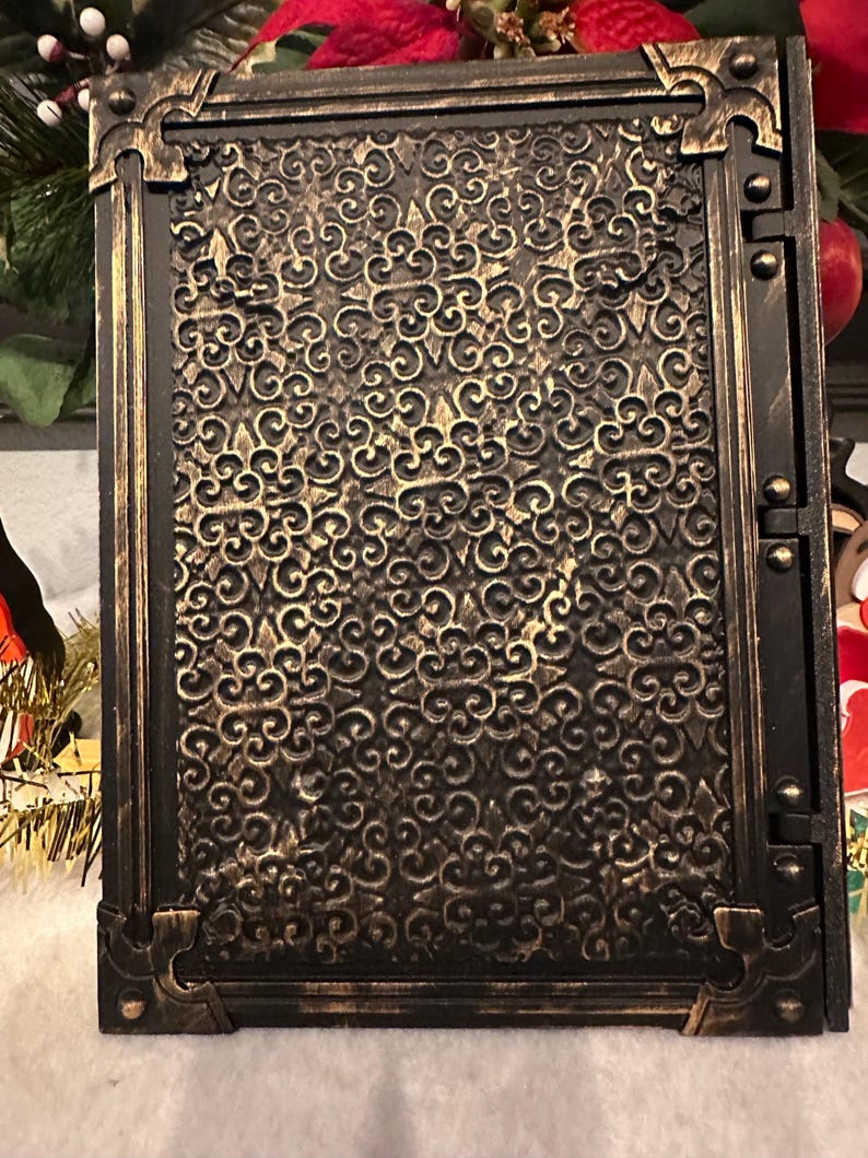 Op de afbeelding: Een rechthoekige, sierlijke, zwart-gouden decoratieve doos. De doos heeft een gedetailleerd, wervelend patroon op het voorpaneel, omlijst door een goudkleurige rand met decoratieve hoekaccenten. De doos heeft een scharnierzijde.