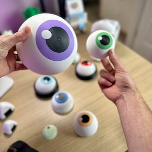 May include: White, spherical novelty eyeballs with various iris colors, including purple, green, blue, and tan. The irises have black pupils. Some eyeballs are held by a person, while others are arranged on a light-colored surface. The eyeballs vary in size.