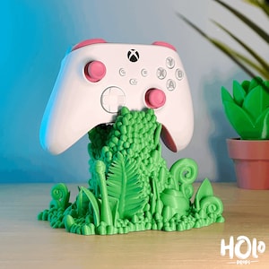 May include: A white video game controller with pink buttons is sitting on a green 3D printed stand. The stand is shaped like a jungle scene with leaves and vines.