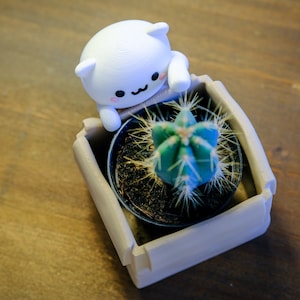 May include: A white ceramic figurine of a cat with a cute face, holding a small brown pot with a green cactus plant inside. The pot is sitting in a larger, light brown planter.