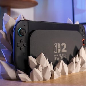 May include: A black Nintendo Switch console rests in a white, geometric crystal-shaped stand. The console has teal and orange accents. The stand is on a wooden surface, with a speaker and a blurred background.