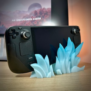 May include: A black handheld gaming device with a blue and white 3D printed stand. The device has a screen, two buttons, and the word "STEAM" printed on it.