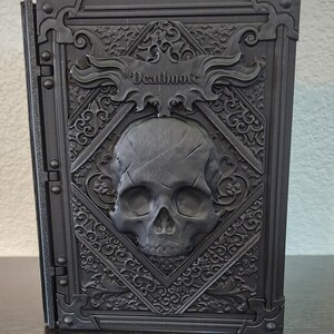 Op de afbeelding: Een zwart, rechthoekig boek met het woord "Deathnote" in reli&euml;f op de kaft. De kaft heeft een schedelontwerp in een ruitvormig kader, omgeven door sierlijke krullen. Het boek staat rechtop.