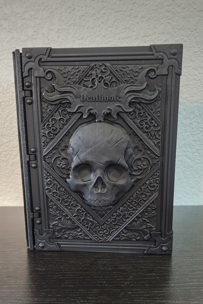 Op de afbeelding: Een zwart, rechthoekig boek met het woord "Deathnote" in reli&euml;f op de kaft. De kaft heeft een schedelontwerp in een ruitvormig kader, omgeven door sierlijke krullen. Het boek staat rechtop.