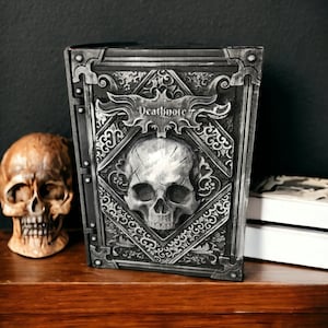 May include: A decorative book with a skull design and the word "Deathnote" on the cover. The book is gray and has intricate detailing. A skull figurine and a stack of books are also visible.