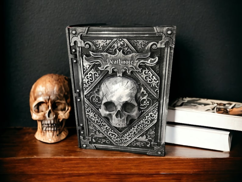 Op de afbeelding: Een decoratief grijs boek met een schedelontwerp en het woord "Deathnote" op de kaft. Het boek heeft ingewikkelde details. Een schedelbeeldje en een stapel boeken zijn ook zichtbaar.