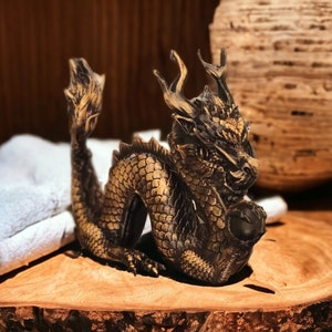 May include: A gold and black dragon figurine with intricate details. The dragon is curled up with its head resting on its front paws. The figurine is sitting on a wooden base.