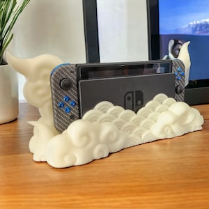 May include: A cream-colored cloud-shaped stand holding a black Nintendo Switch console. The stand has a detailed cloud design, and the console features blue buttons and a carbon fiber pattern on the controllers. The background includes a TV screen.