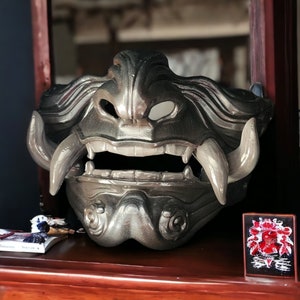 May include: A silver and gray Oni mask with a fierce expression. The mask has large, sharp teeth and a wide, open mouth. The mask is made of a hard, durable material and has a detailed design.