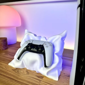 May include: A white PlayStation 5 controller resting on a white pillow with a blue light shining from underneath. The controller has black buttons and a white and black design.