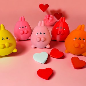 May include: Five colorful bunny figurines with hearts scattered around them. The bunnies are pink, yellow, orange, and red. The hearts are red, pink, and light blue.