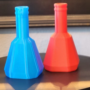 May include: Two decorative glass bottles, one blue and one red, with a diamond-shaped base and a narrow neck. The bottles are sitting on a black surface.