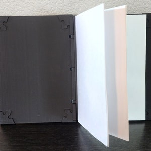Op de afbeelding: Een zwart, open boek met witte pagina's. De kaft en achterkant zijn zwart met een textuur en hoekontwerpen in de vorm van puzzelstukjes. De pagina's zijn wit en lichtgroen.