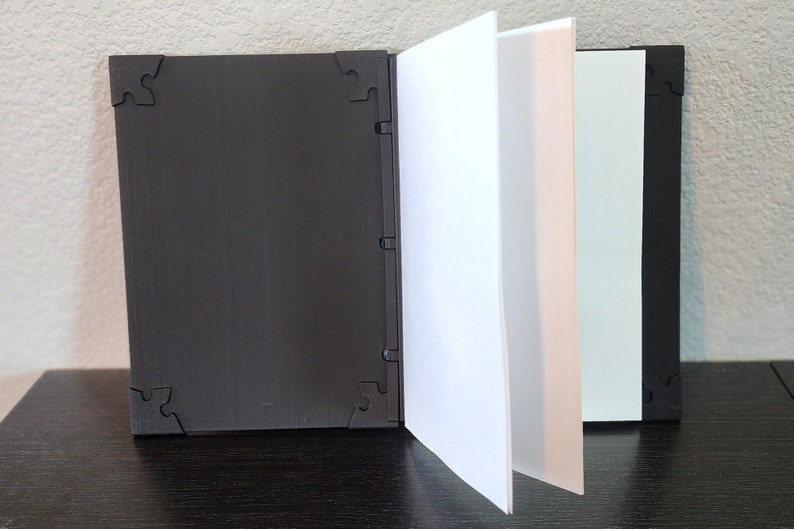 Op de afbeelding: Een zwart, open boek met witte pagina's. De kaft en achterkant zijn zwart met een textuur en hoekontwerpen in de vorm van puzzelstukjes. De pagina's zijn wit en lichtgroen.