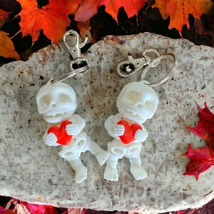 May include: Two white skeleton keychains, each holding a red heart. The keychains have silver clasps and are set against a stone surface with colorful autumn leaves in the background. A unique, spooky, and romantic accessory.