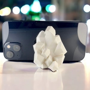 May include: A white, 3D printed, crystal-shaped phone stand. The stand is made to look like a cluster of crystals.