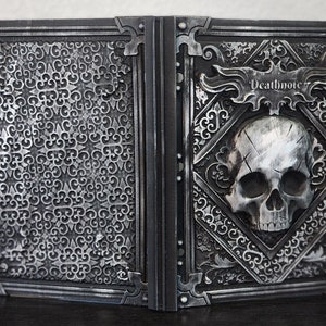 Op de afbeelding: Een open boek met een gedetailleerd zilverkleurig ontwerp. De rechterkant toont een schedel en het woord "Deathnote". De linkerkant heeft een zich herhalend patroon. Het boek is waarschijnlijk een decoratief item of rekwisiet.