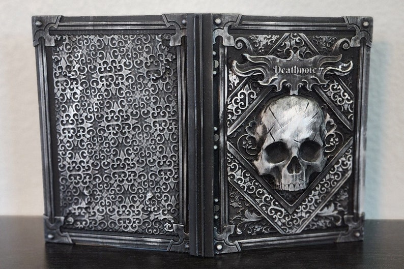 Op de afbeelding: Een open boek met een gedetailleerd zilverkleurig ontwerp. De rechterkant toont een schedel en het woord "Deathnote". De linkerkant heeft een zich herhalend patroon. Het boek is waarschijnlijk een decoratief item of rekwisiet.