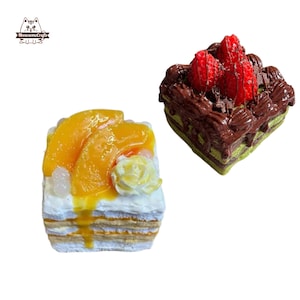 Puede incluir: Dos pasteles en miniatura. Uno tiene capas blancas y azules con rodajas de melocotón y una flor amarilla. El otro tiene una base verde, glaseado de chocolate y fresas encima. Los pasteles están sobre un fondo blanco.