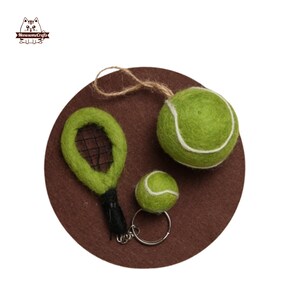 May include: A felt tennis-themed set featuring a green tennis ball, a miniature tennis racket, and a tennis ball keychain. The items are arranged on a brown circular base with a natural twine string attached to the ball.