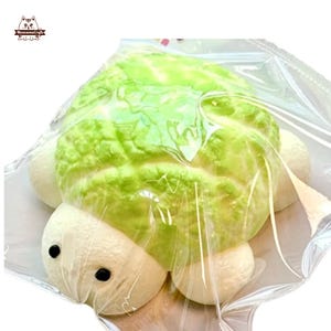 May include: A handmade turtle-shaped pastry, with a green top and white body parts, is wrapped in clear plastic. The turtle has two black dot eyes. The pastry is a unique food item.