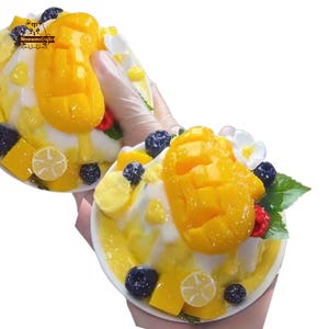 May include: Two bowls of dessert are held in the image. Each bowl is filled with a yellow, creamy base, topped with large pieces of yellow mango, blueberries, raspberries, and lemon slices. The bowls are garnished with white flowers and green leaves. The text "Meowsome Crafts" is visible.