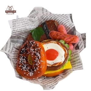 May include: A colorful assortment of miniature food items, including a burger with a fried egg, sausages, and a bagel with sesame seeds, arranged on a paper-lined tray. The food items are brightly colored and appear to be made of resin or a similar material.