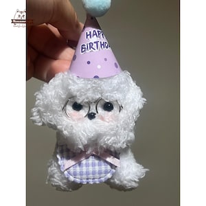 May include: A white plush dog toy wearing a purple party hat with the words "Happy Birthday" and a gingham bib. The dog has black eyes and wire-rimmed glasses. A blue pom-pom is on top of the hat.