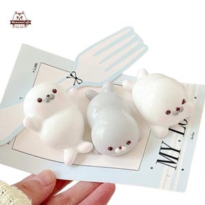 以下が含まれることがあります： 白とグレーの可愛らしいアザラシの置物3体。顔には小さな赤いアクセントがあります。「MY Love」の文字とフォークのデザインが施された水色のカードの上に配置されています。手作りの置物です。