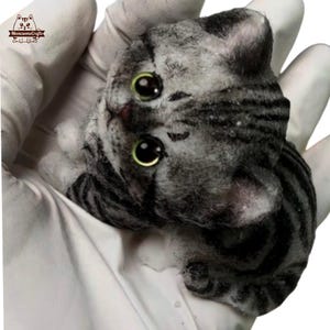 Puede incluir: Una pequeña figurita de gato rayada gris y blanca con grandes ojos verdes. El gato está acostado de espaldas con las patas en el aire.