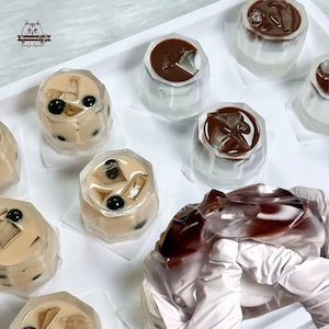May include: A tray of clear plastic cups filled with brown and white liquid, some with black boba balls. The cups are arranged in a grid pattern. There is a hand holding a clear plastic cup filled with brown liquid.