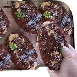 Può includere: Primo piano di diversi pasticcini al cioccolato con un ricco colore marrone scuro. Ogni pasticcino è ricoperto da una crema colorata chiara, scaglie di cioccolato e un rametto verde. Piccole etichette con la scritta "Happy Everyday".