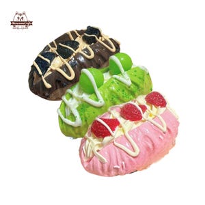 Può includere: Tre pasticcini decorativi oblunghi di diversi colori. Il pasticcino superiore è al cioccolato con biscotti e glassa bianca. Il pasticcino centrale è verde con palline verdi e glassa bianca. Il pasticcino inferiore è rosa con fragole e glassa bianca.