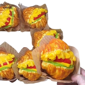 May include: Several croissant sandwiches with yellow, red, and green fillings are displayed. The sandwiches are wrapped in brown paper and appear to be a food item. The image is well-lit, showcasing the details of the food.