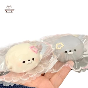 Puede incluir: Dos adorables figuras con forma de foca, una de color crema con un lazo rosa y la otra gris con una estrella amarilla, están empaquetadas en plástico transparente. Las focas tienen ojos negros y rasgos pequeños. Las figuras se sostienen en una mano.