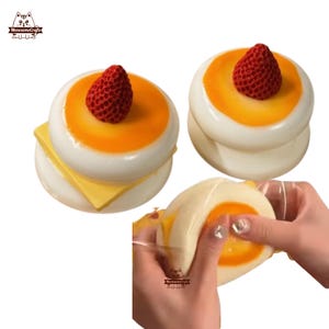 May include: Two white squishy toys shaped like a Japanese steamed bun with a yellow custard filling and a red strawberry on top. One bun is stacked on top of another. The third bun is being squeezed by a hand.