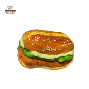 Può includere: Un hamburger artigianale dettagliato, con un panino ai semi di sesamo, un patty marrone, relish verde e una salsa cremosa. L'hamburger è una replica in miniatura, probabilmente un oggetto decorativo o un artigianato a tema alimentare.