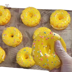 May include: A collection of yellow glazed donuts with colorful sprinkles. The donuts are arranged on a newspaper-printed surface. One donut is being held by a gloved hand. The donuts have a hole in the center.