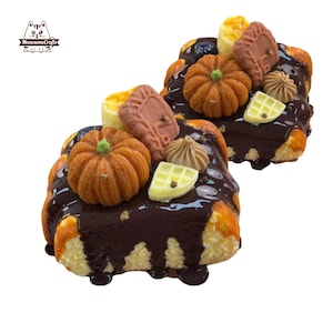 Può includere: Due pasticcini in miniatura con glassa al cioccolato, guarniti con decorazioni di zucca e biscotti. I pasticcini hanno una base marrone chiaro e sono decorati con zucche arancioni, biscotti marroni chiari e caramelle bianche a forma di waffle.