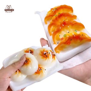 Può includere: Due piatti di repliche di cibo in miniatura. Un piatto contiene quattro panini bianchi e rotondi con dettagli arancioni e neri. L'altro piatto contiene quattro ravioli arancioni con accenti rossi e verdi. Entrambi i piatti sono bianchi.